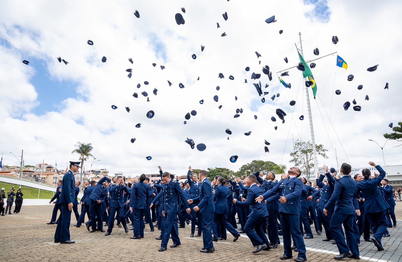 Turma de cadetes comemorando a formatura, jogando os quepes para o alto em frente ao mastro com a bandeira do Brasil